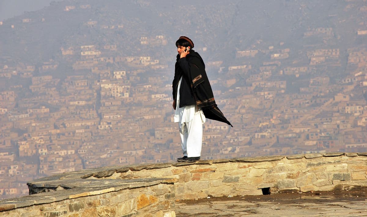 A man stands on a wall with a city behind him, using a mobile phone.
