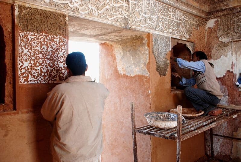 Men work on partially restored orange walls with calligraphy and floral designs.